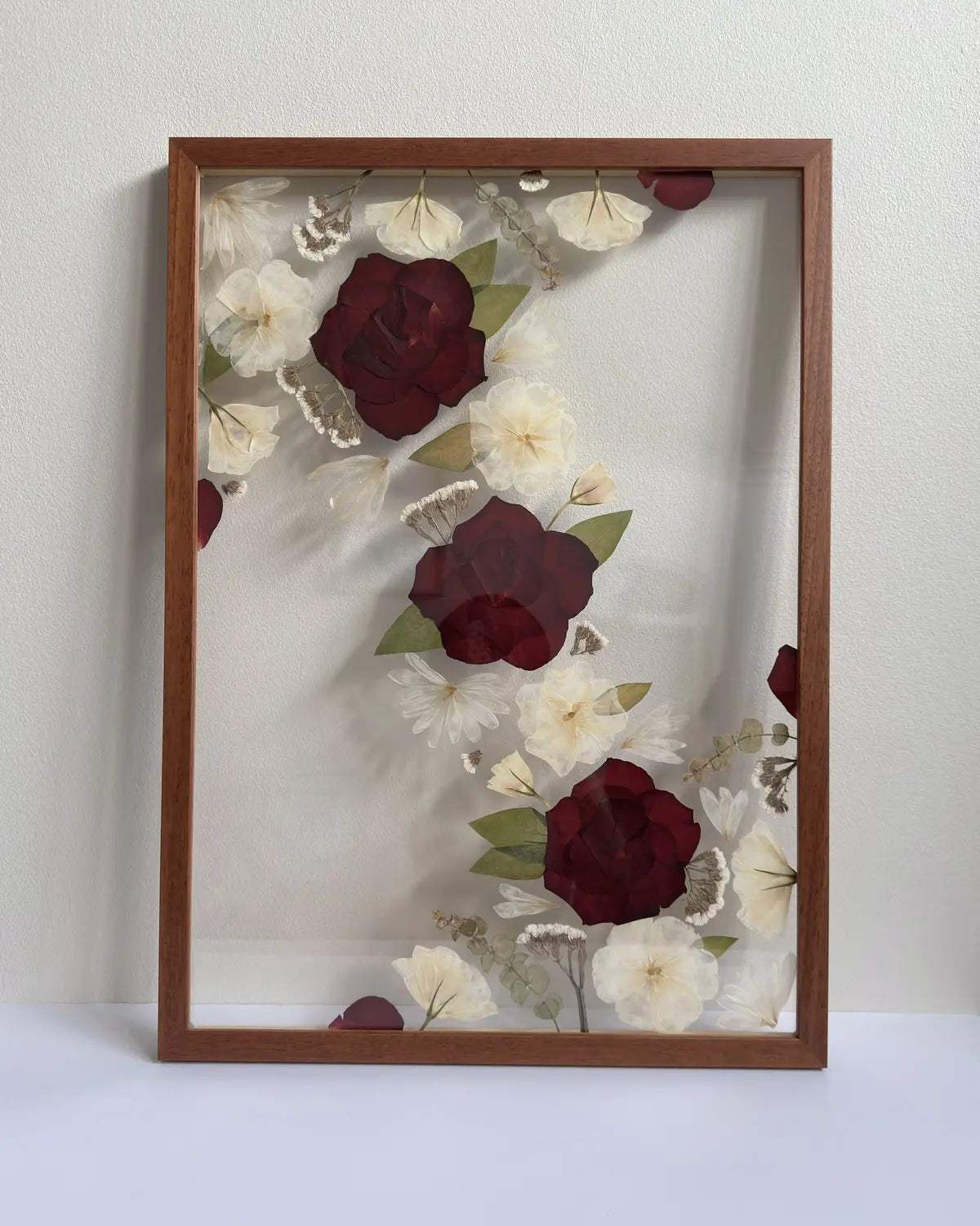 Framed artwork with red and white pressed flowers on a plain background in a warm oak frame