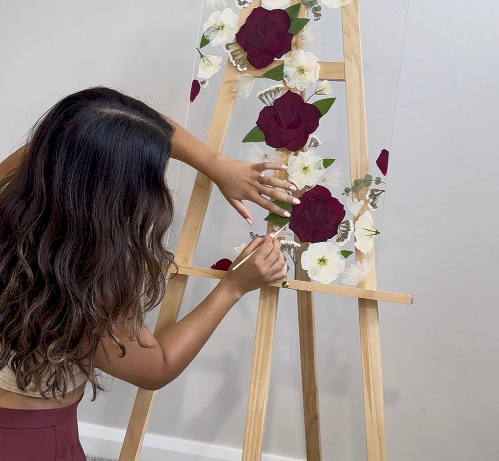 Person arranging pressed flowers on a wooden easel against a plain wall.