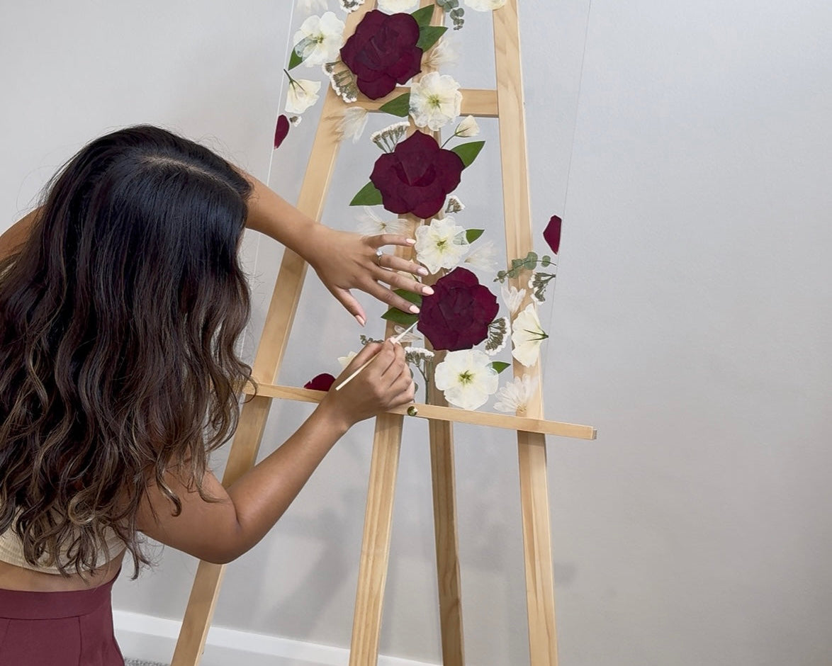Person arranging pressed flowers on a wooden easel against a plain wall.