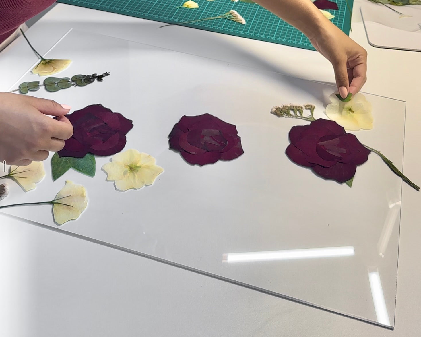 Person arranging pressed flowers on a white surface with a green cutting mat in the background.