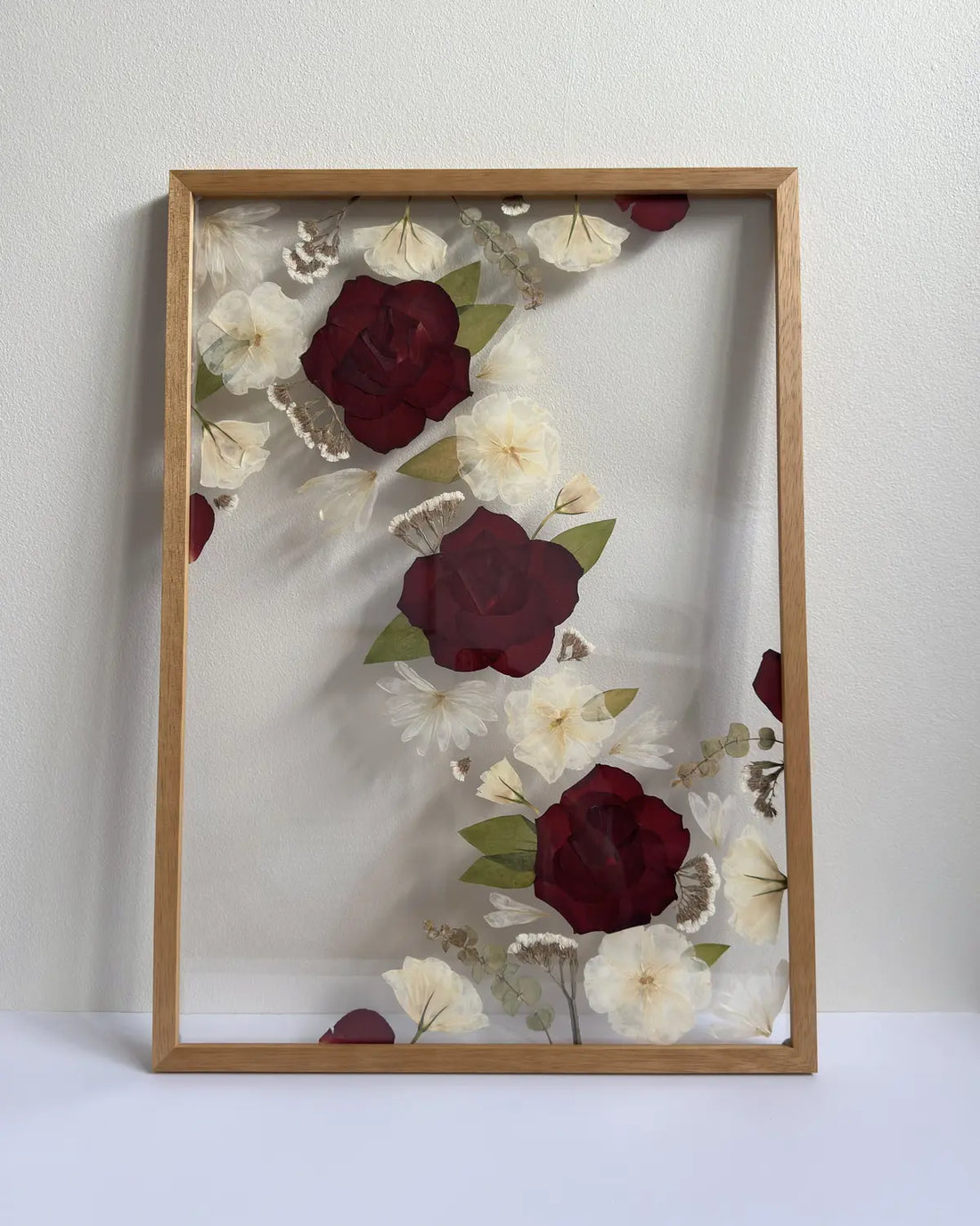 Framed artwork with pressed flowers including red and white roses in a light, natural oak coloured frame
