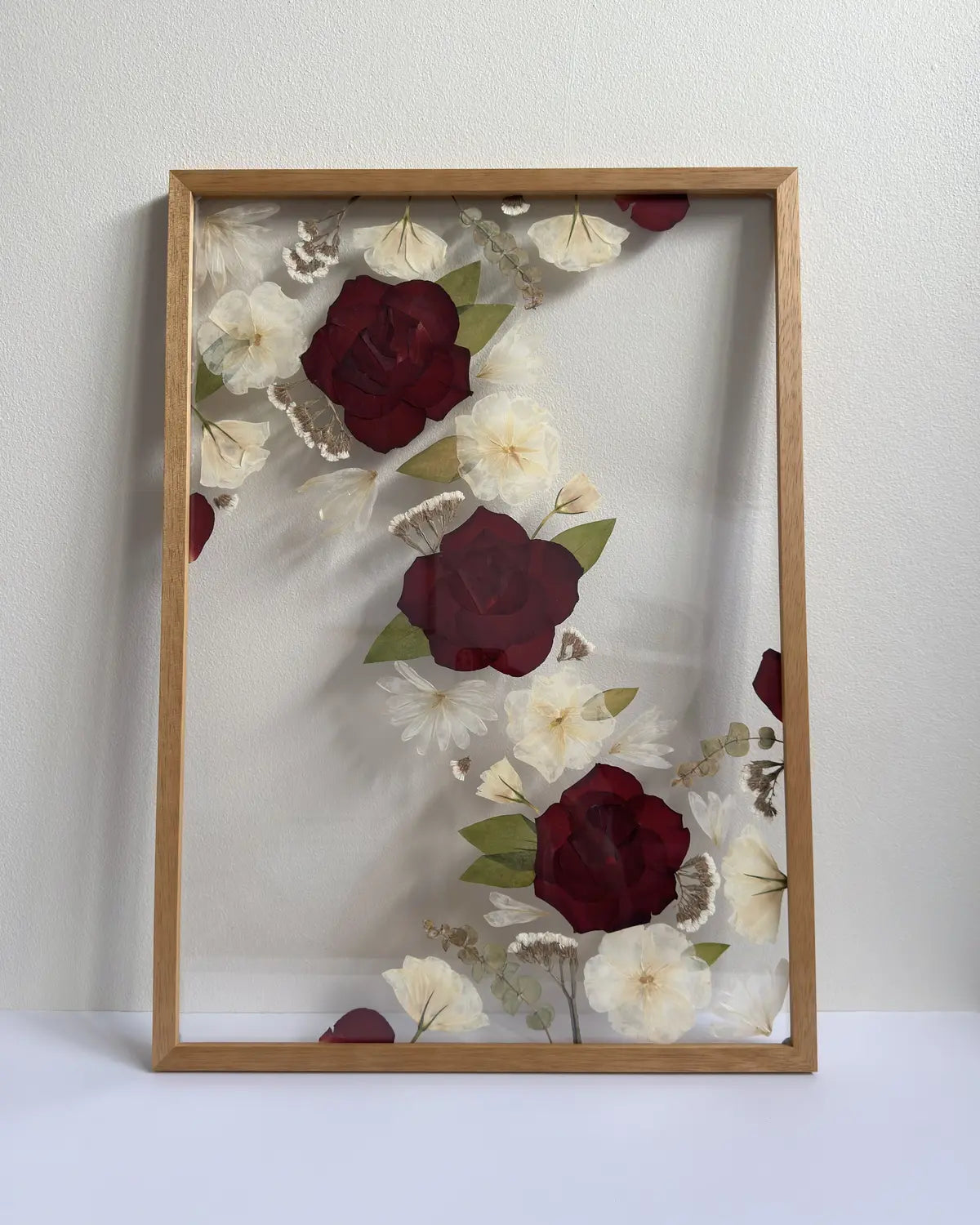 Framed artwork with pressed flowers including red and white roses in a light, natural oak coloured frame