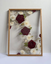 Framed artwork with pressed flowers including red and white roses in a light, natural oak coloured frame